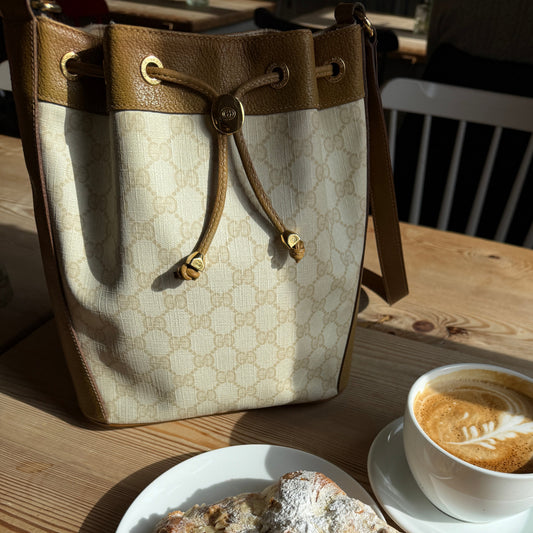 Café setting with a pastry on a plate and a cup of coffee on a saucer, next to a Gucci handbag on a wooden table.